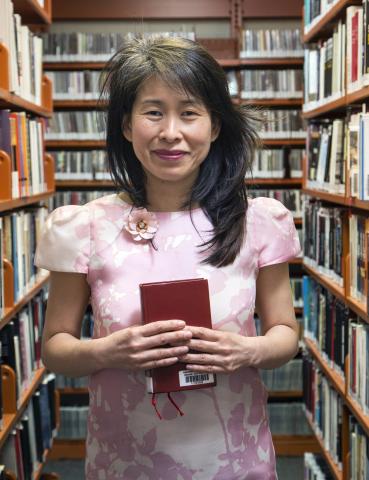 Kim Thúy, avec un livre à la main, portant une robe rose et entourée d'étagères.