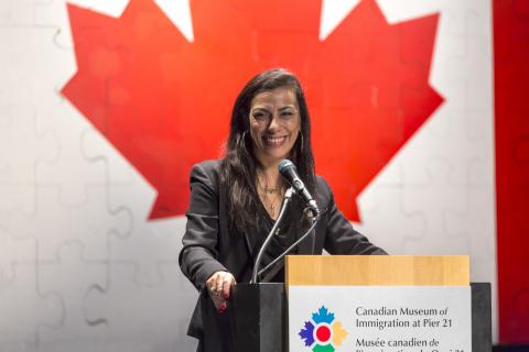 Carmen Aguirre à la tribune, devant un grand drapeau canadien.