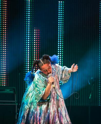 Lido Pimienta sur scène dans une robe multicolore et les cheveux tressés.