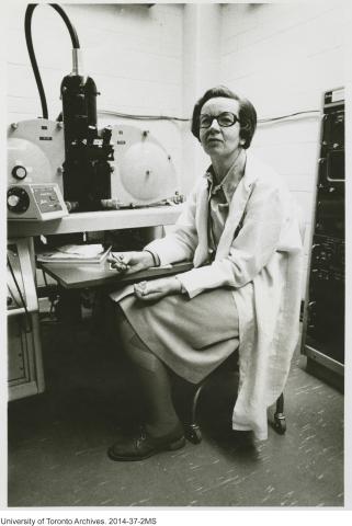 Photo en noir et blanc d’un scientifique en blouse de laboratoire, assis devant un gros équipement de laboratoire.
