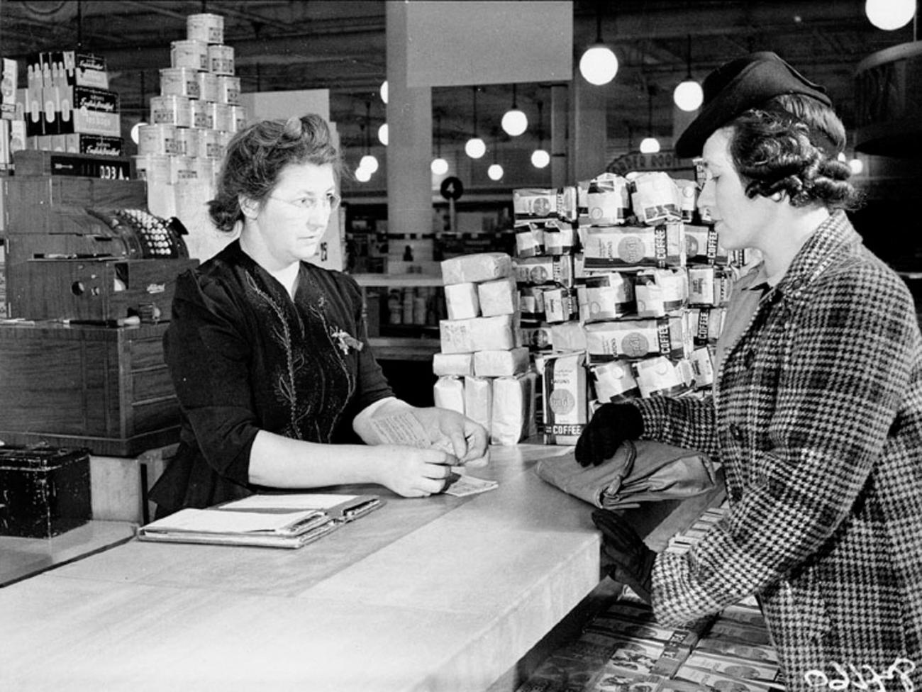 Deux femmes discutent à un comptoir de magasin avec des piles de produits derrière elles.