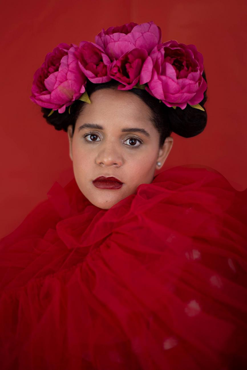 Photo de la tête et des épaules de Lido Pimienta, le corps recouvert d’un filet rouge, les cheveux relevés avec de grosses fleurs roses.