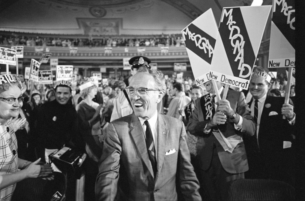 Photo en noir et blanc de Tommy Douglas dans un théâtre avec des personnes derrière lui brandissant des pancartes.