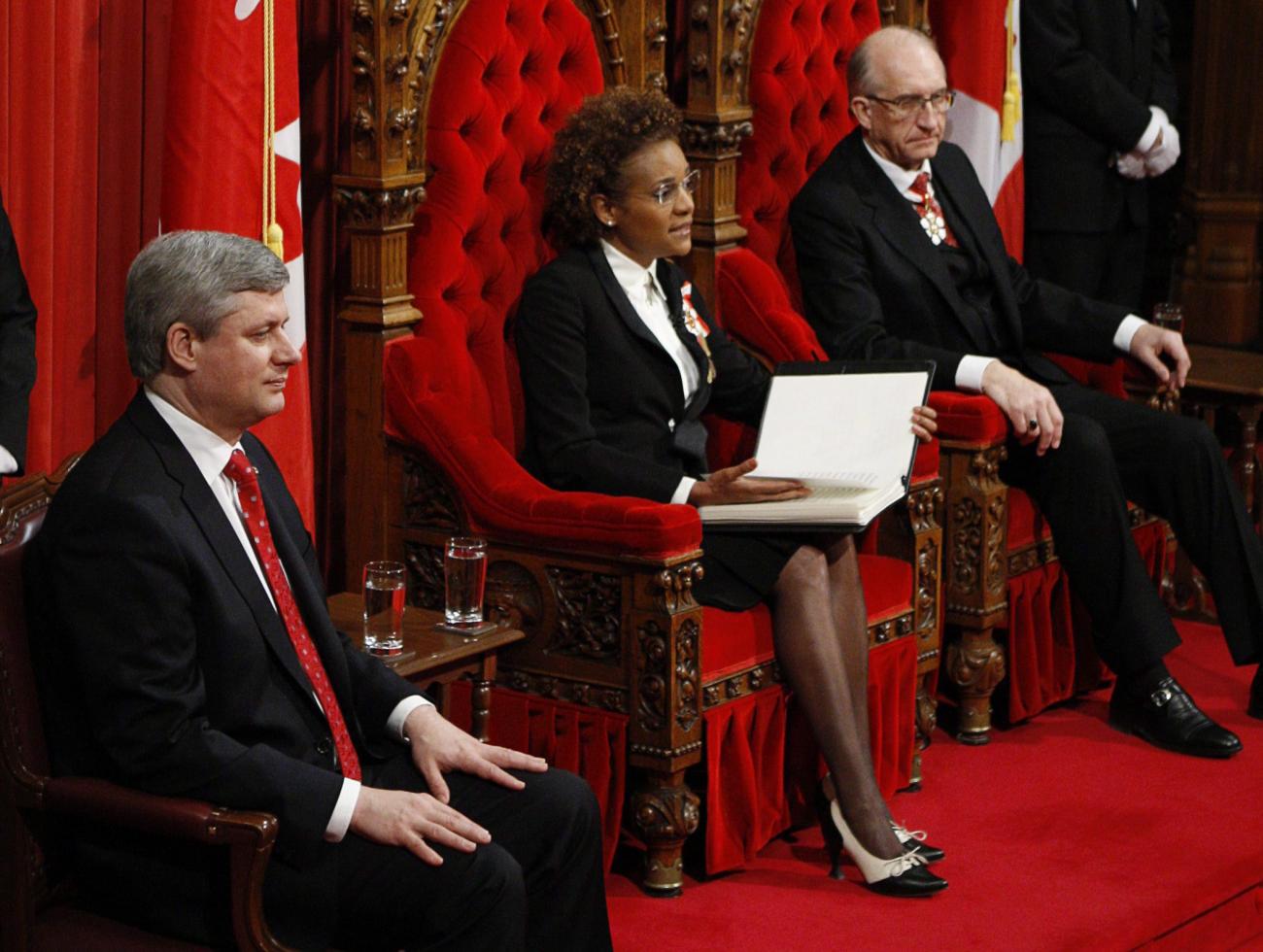 Michaëlle Jean assise sur un trône rouge entre deux hommes, l’un sur un trône rouge, l’autre sur une chaise.