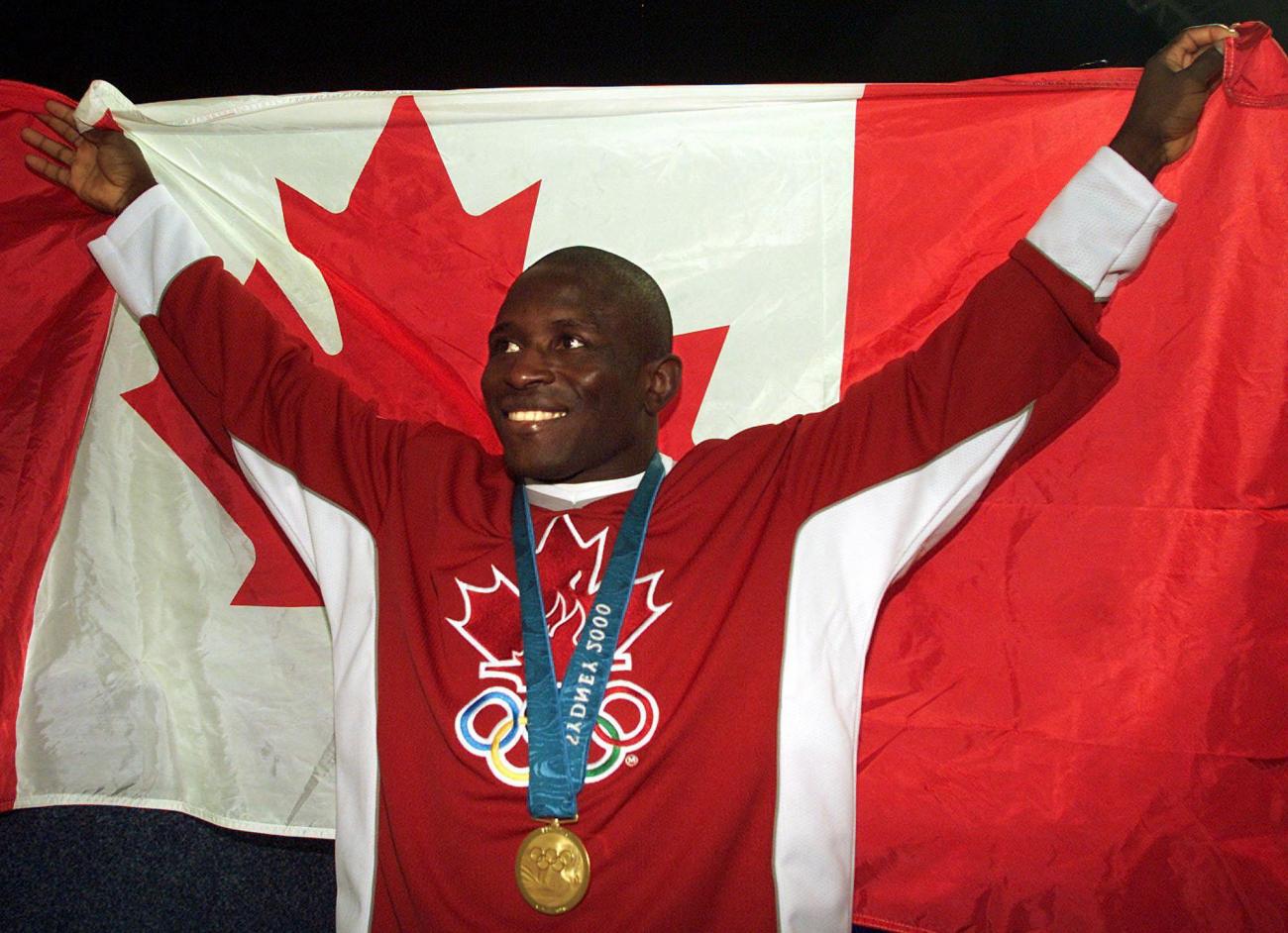 Daniel Igali, vêtu de rouge et portant une médaille, tient un drapeau canadien derrière lui.