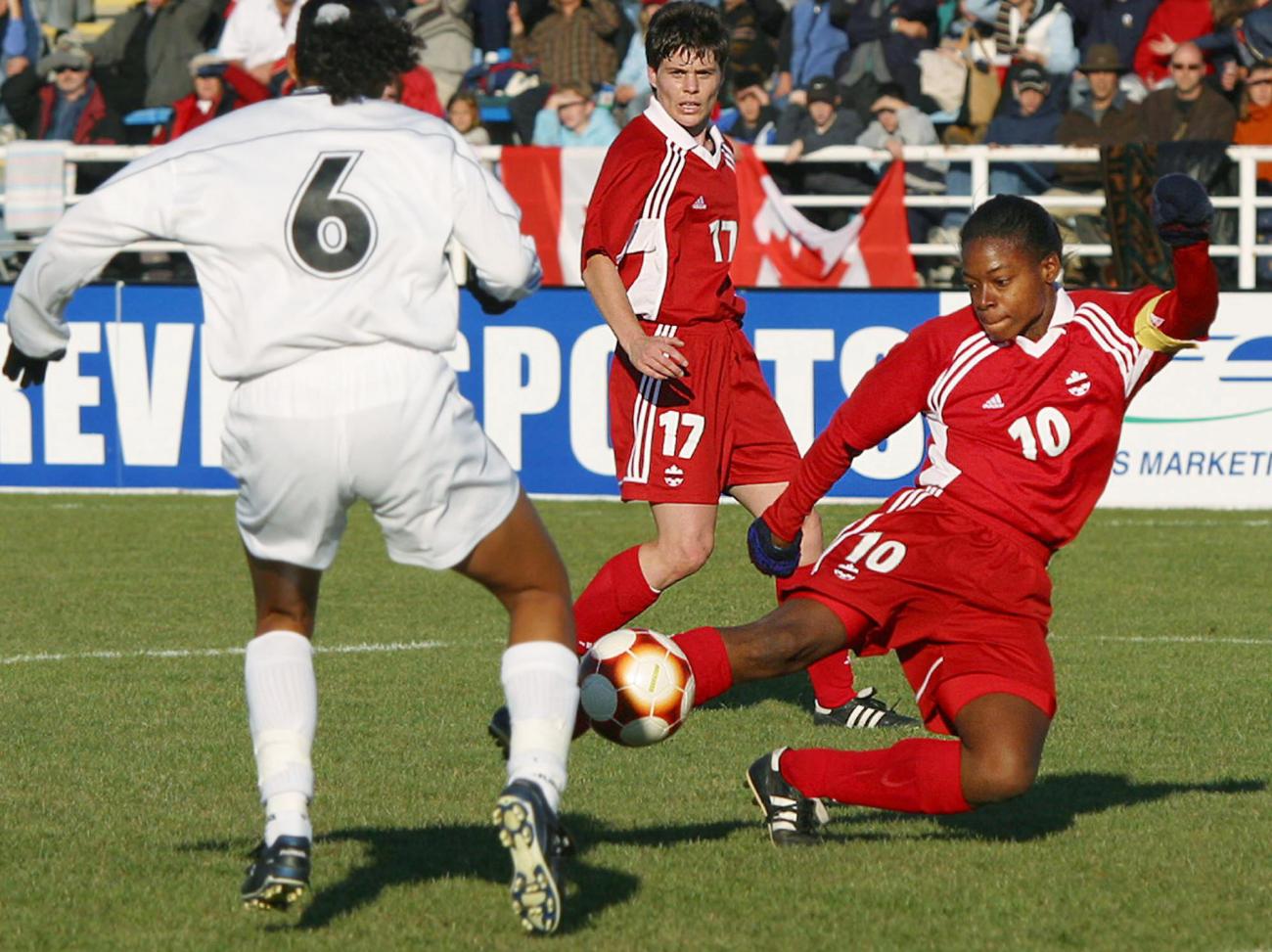 Charmaine Hooper en rouge sur un terrain de soccer avec deux autres joueuses, l’une en rouge, l’autre en blanc.