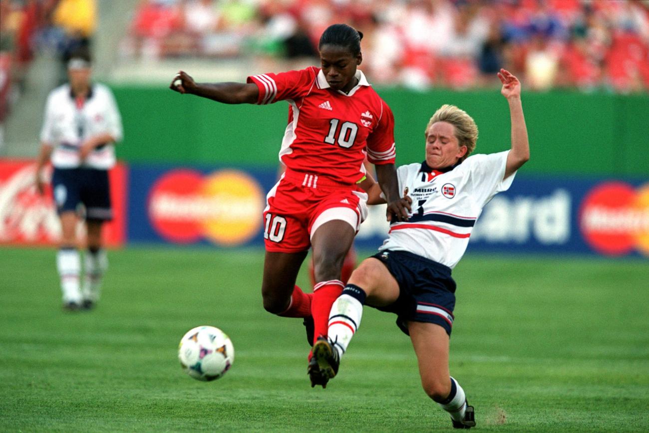Charmaine Hooper en rouge et une autre joueuse en blanc sur un terrain de soccer en train de courir après le ballon.