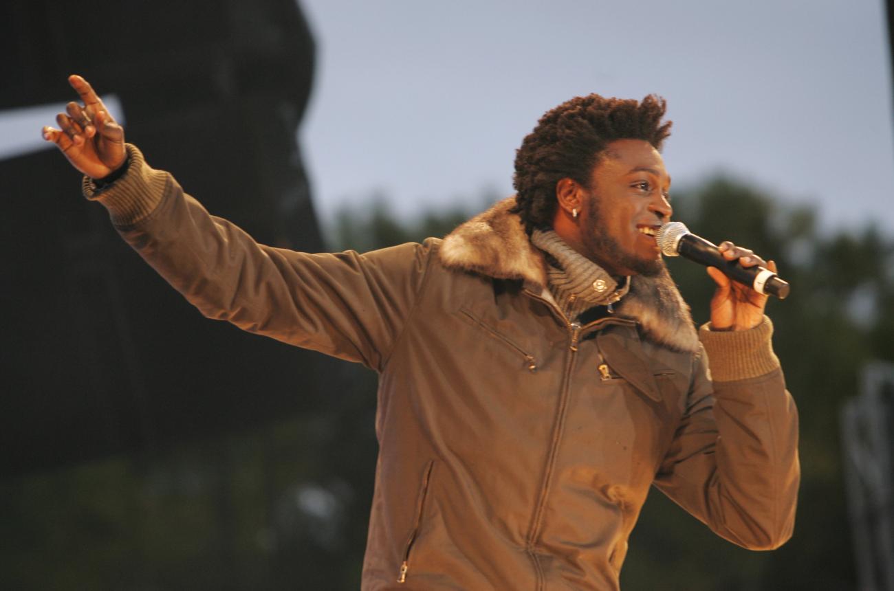 Corneille sur scène en plein air, chantant le bras tendu et portant un manteau brun.
