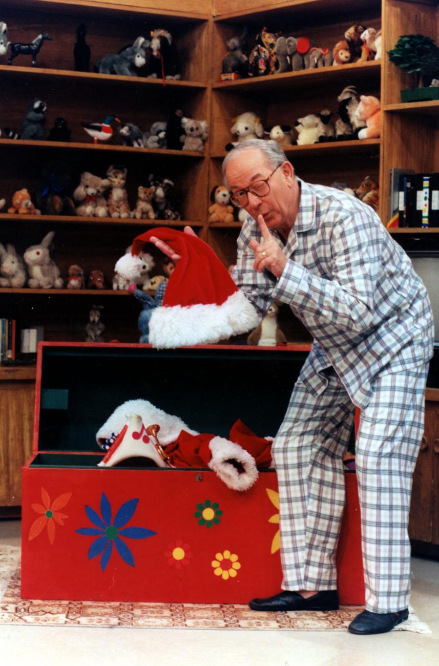Ernie Coombs dans le rôle de Mr. Dressup en pyjama tirant un chapeau de Père Noël d’une malle dans une pièce bordée d’étagères de jouets.
