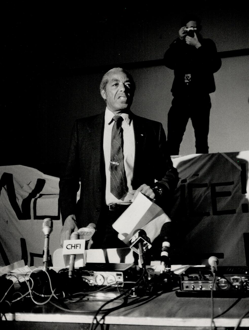 Bromley Armstrong, en costume et cravate, un papier à la main, se tient debout avec plusieurs microphones devant lui.