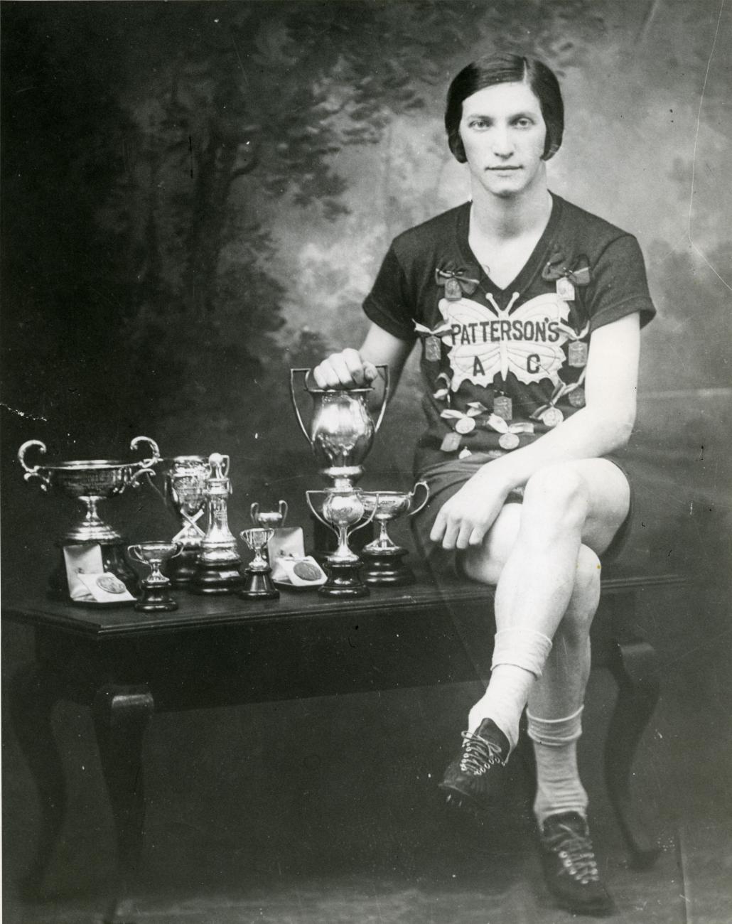 Bobbie Rosenfeld assise sur une petite table à côté de nombreux trophées.