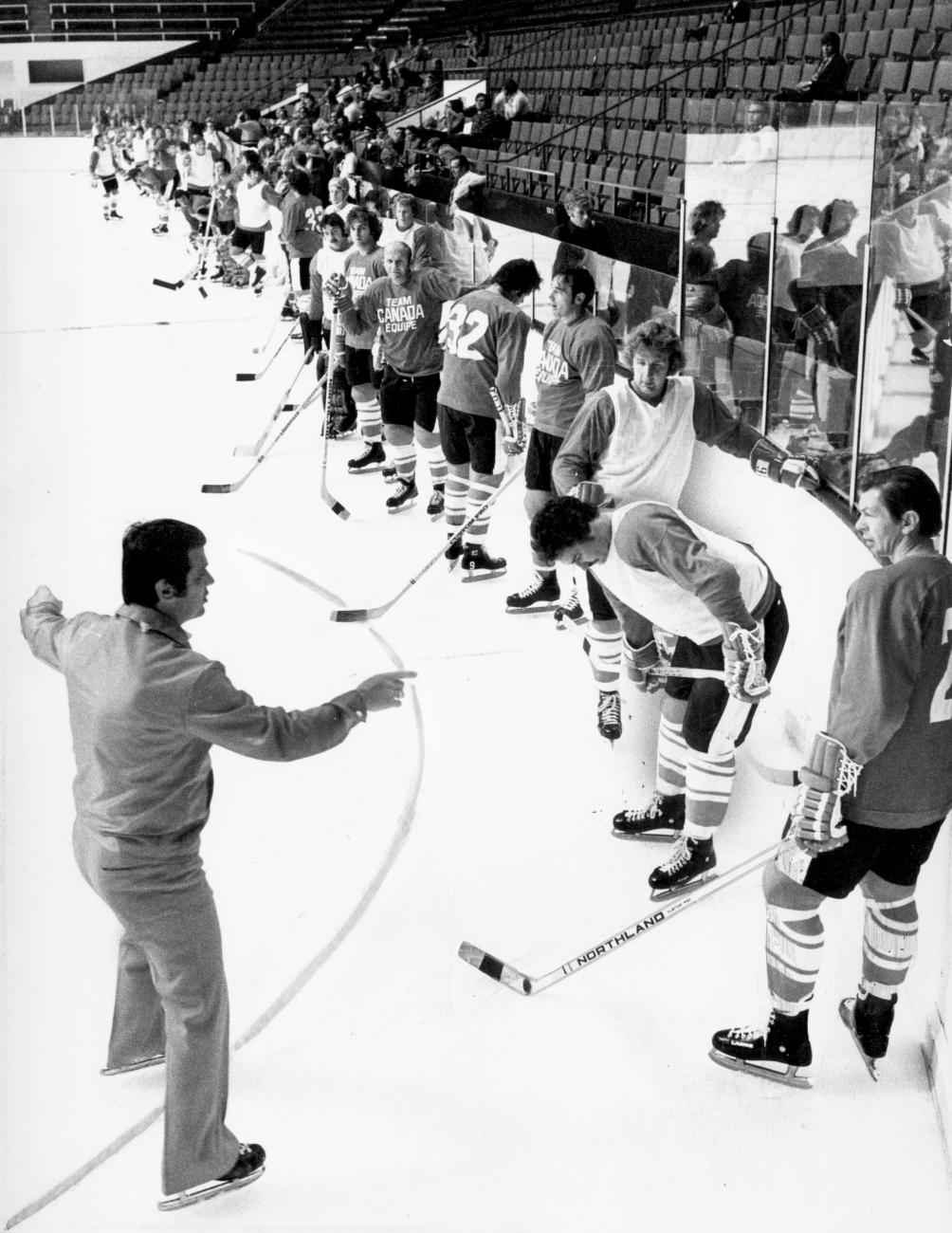 Une ligne de joueurs de hockey le long des bandes de la patinoire avec Stan Mikita parlant à l’entraîneur au premier plan.