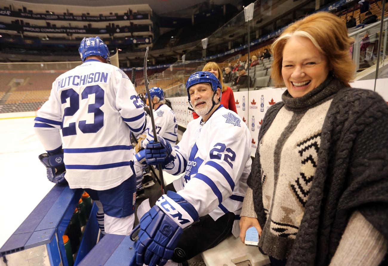 Une femme sourit pour la caméra, debout dans la zone des entraîneurs, derrière un banc de hockey avec des joueurs de hockey à la retraite portant des uniformes des Toronto Maple Leaf.