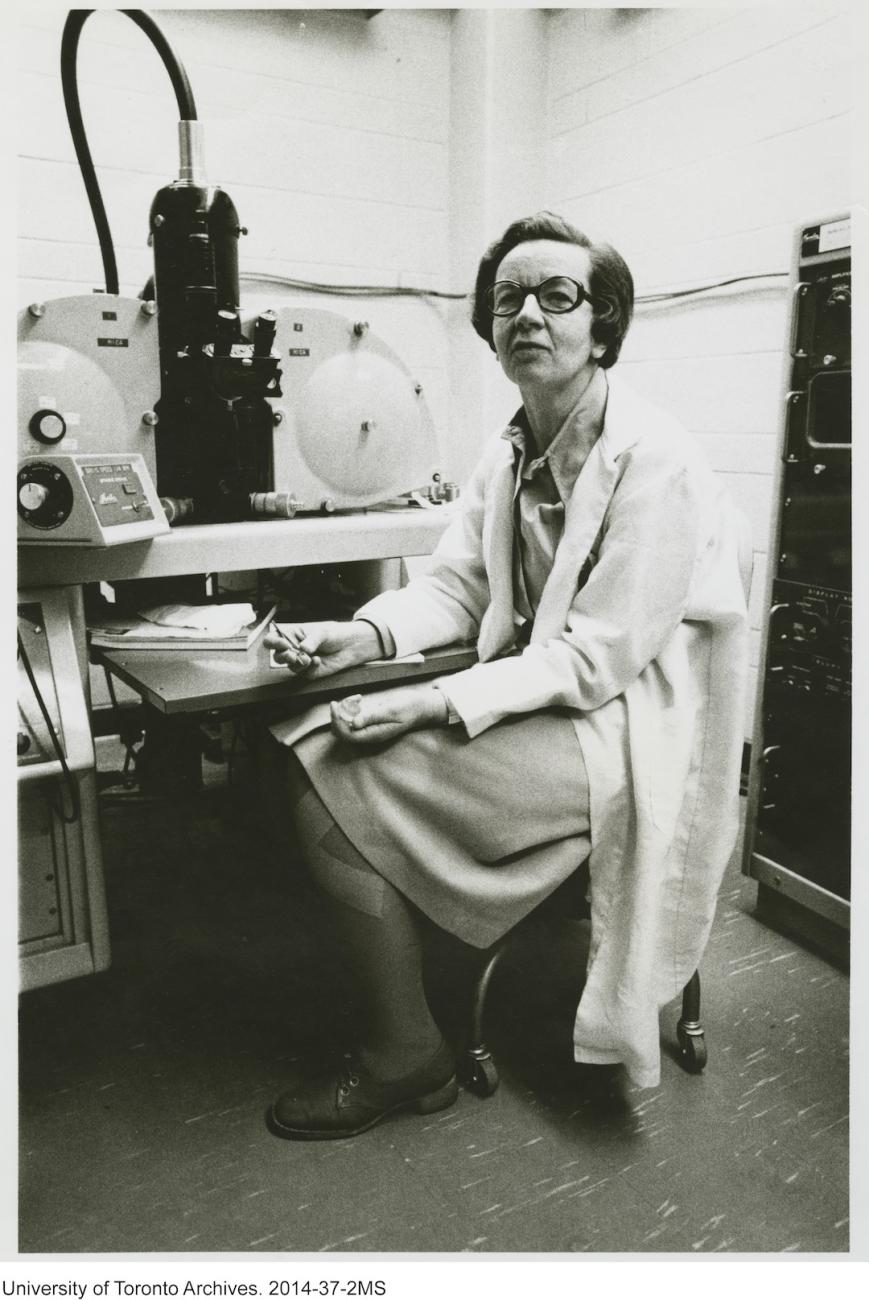 Photo en noir et blanc d’un scientifique en blouse de laboratoire, assis devant une grande pièce d’équipement de laboratoire.