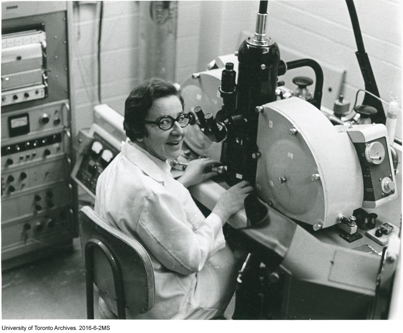 Une scientifique sourit à la caméra depuis sa place devant un microscope.